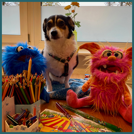 Schulhund Ella mit den Maskottchen Frieda, Fridolin. Viele Buntstifte und ein Zirkusbild auf einem Schreibtisch.