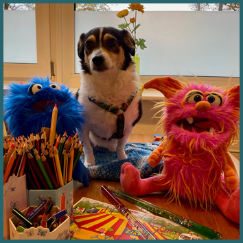 Schulhund Ella mit den Maskottchen Frieda, Fridolin. Viele Buntstifte und ein Zirkusbild auf einem Schreibtisch.